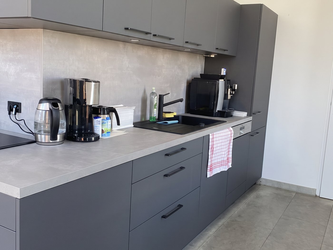 Graue Büroküche mit Spüle, Kaffeemaschine und Wasserkocher auf heller Arbeitsplatte